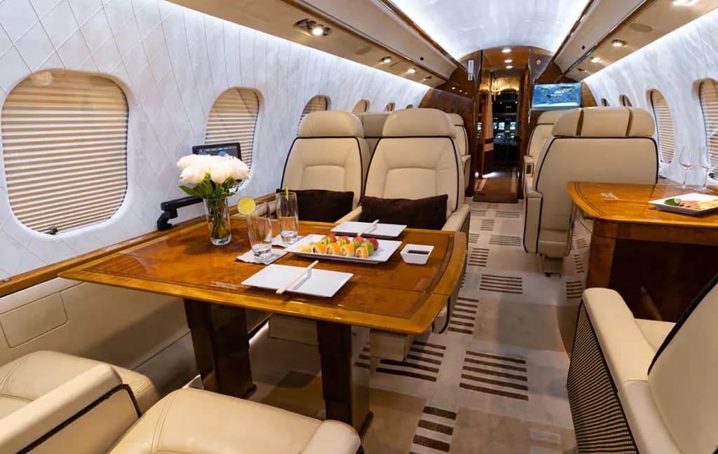 The spacious private jet cabin featuring multiple beige seats, a large wooden table set with sushi and white flowers.