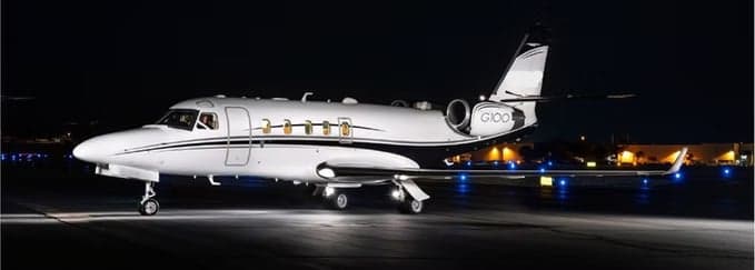 A white Gulfstream G100 business jet with black and gold stripes is shown on a runway at night, illuminated by ground lights.