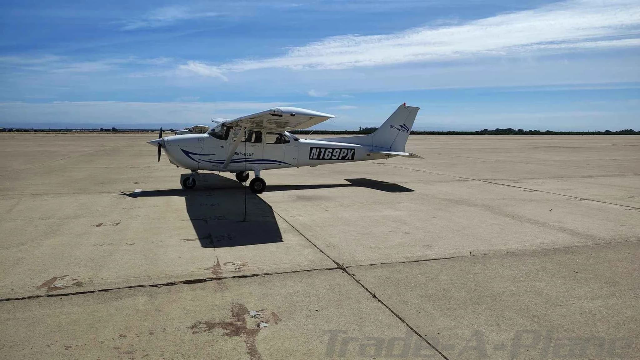 A white Cessna 172 with blue stripes, registration N169PX, parked on an airfield under a blue sky.