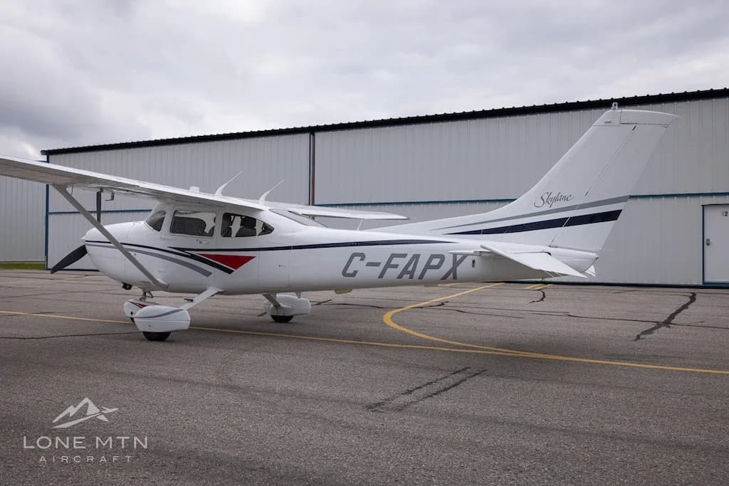A white Cessna 182 Skylane with grey and blue stripes, registration C-FAPX, parked on an asphalt ramp with hangars in the background.