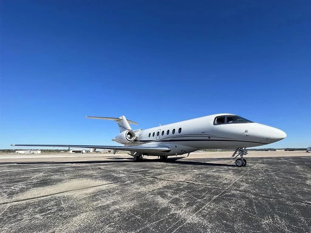 A white business jet with dark blue stripes is parked on an asphalt tarmac, viewed from the front-right.