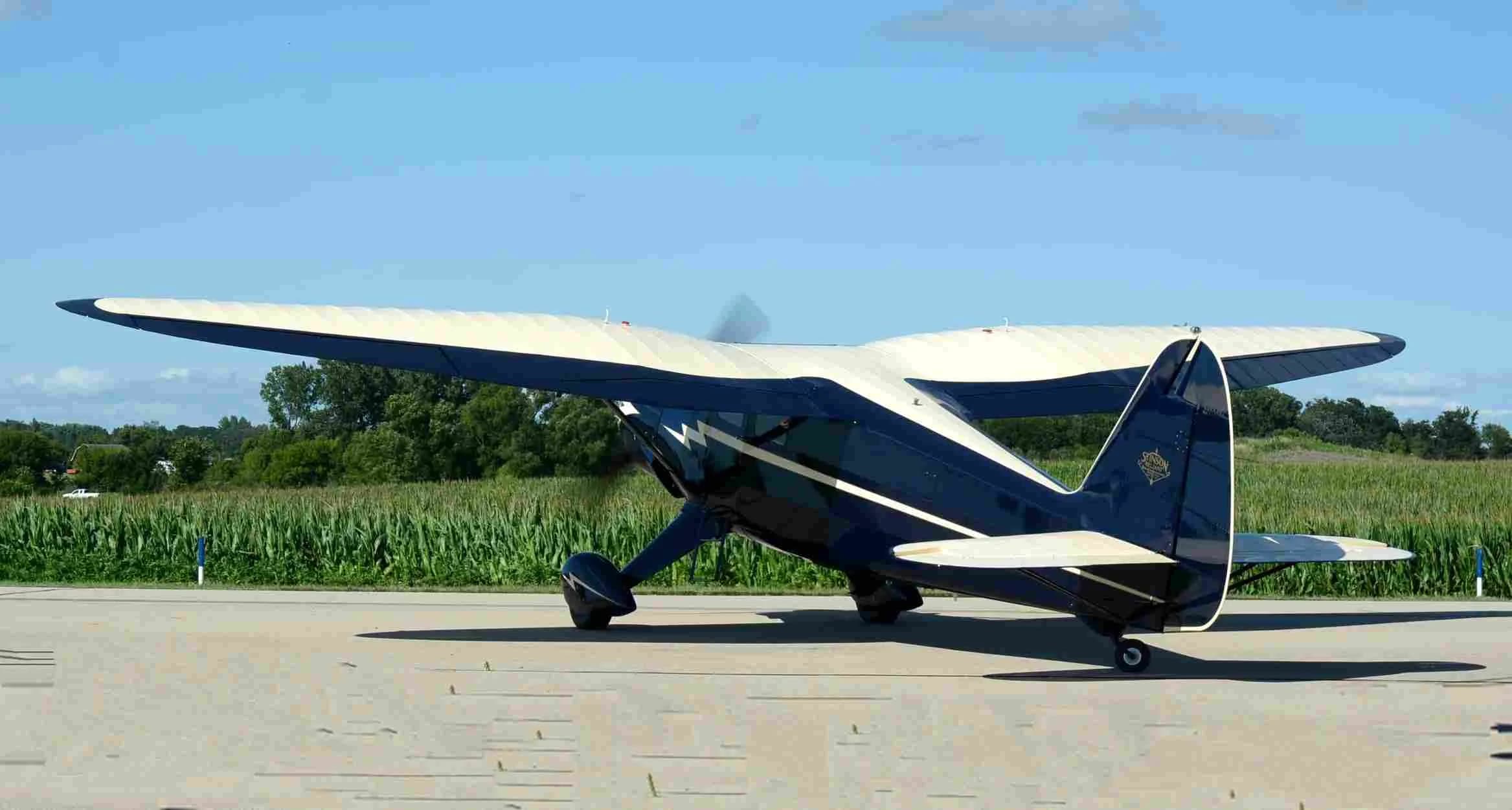 A dark blue and cream vintage aircraft with a lightning bolt design on its fuselage is parked on a tarmac next to a green field.