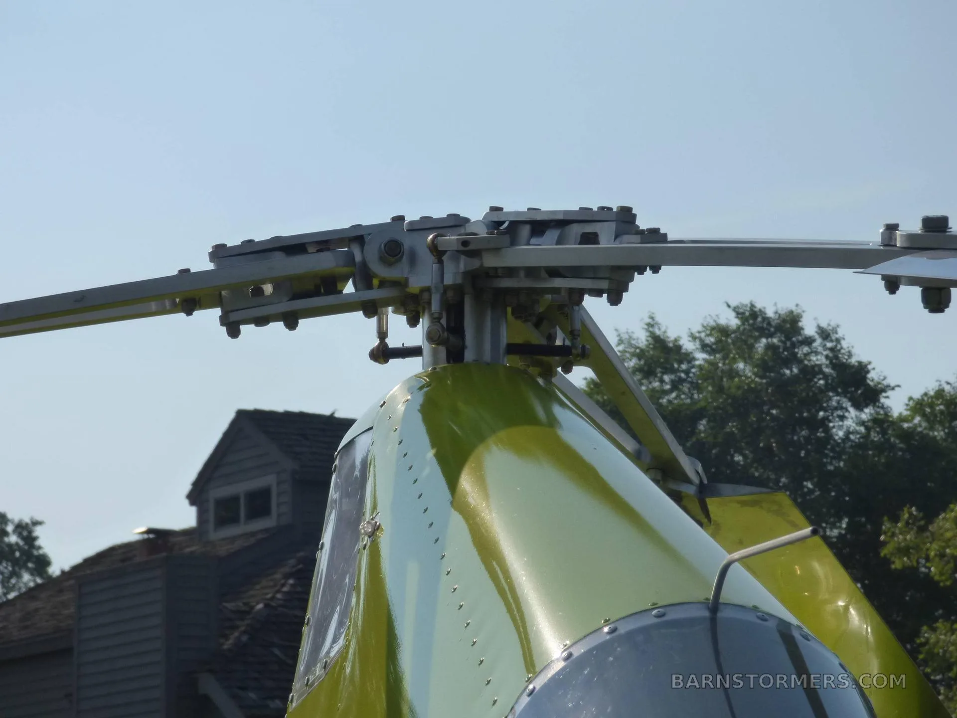 A close-up view of the main rotor head and the top of the bright green gyrocopter's fuselage, with a building and trees in the background.