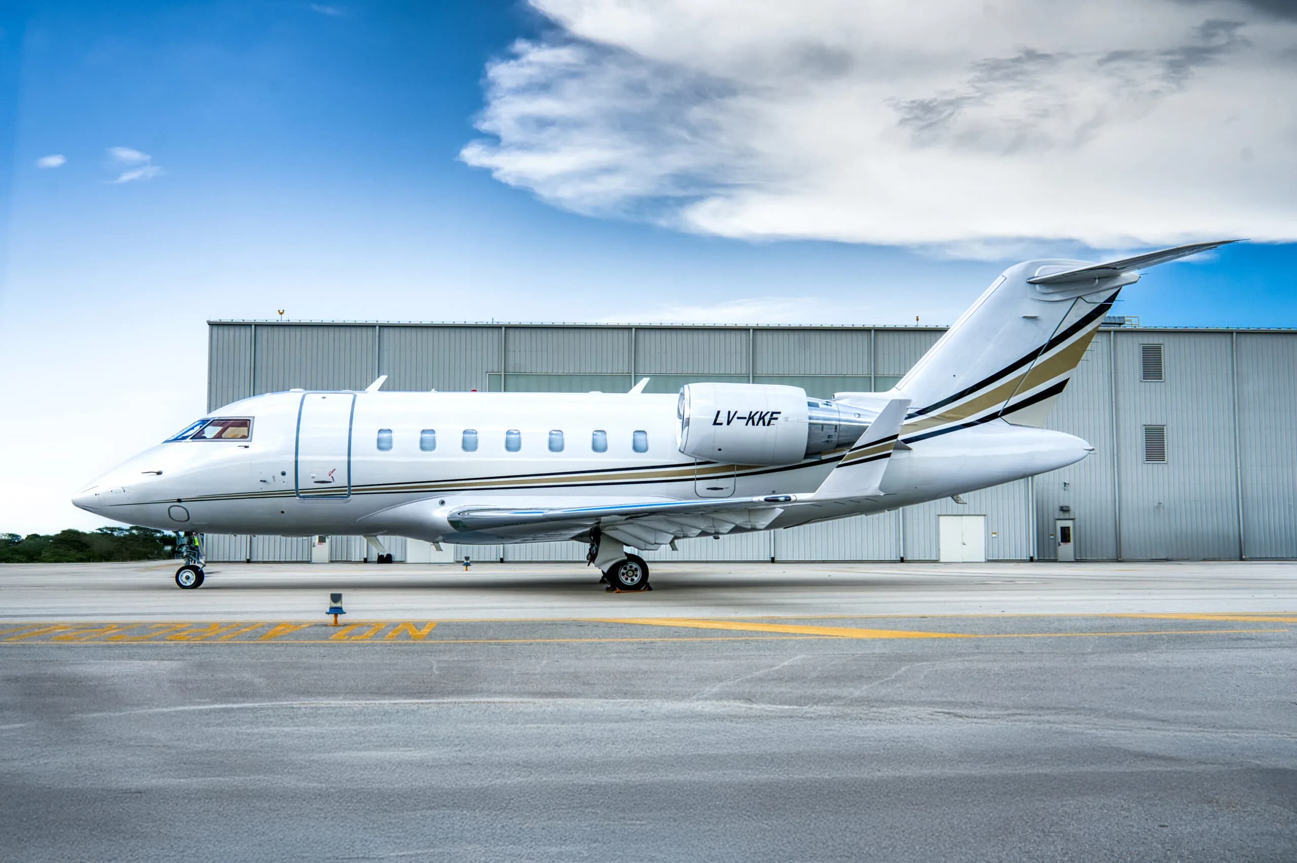 A white Bombardier Challenger business jet with gold and black stripes, registration LV-KKF, seen from the side on an airport tarmac next to a hangar.