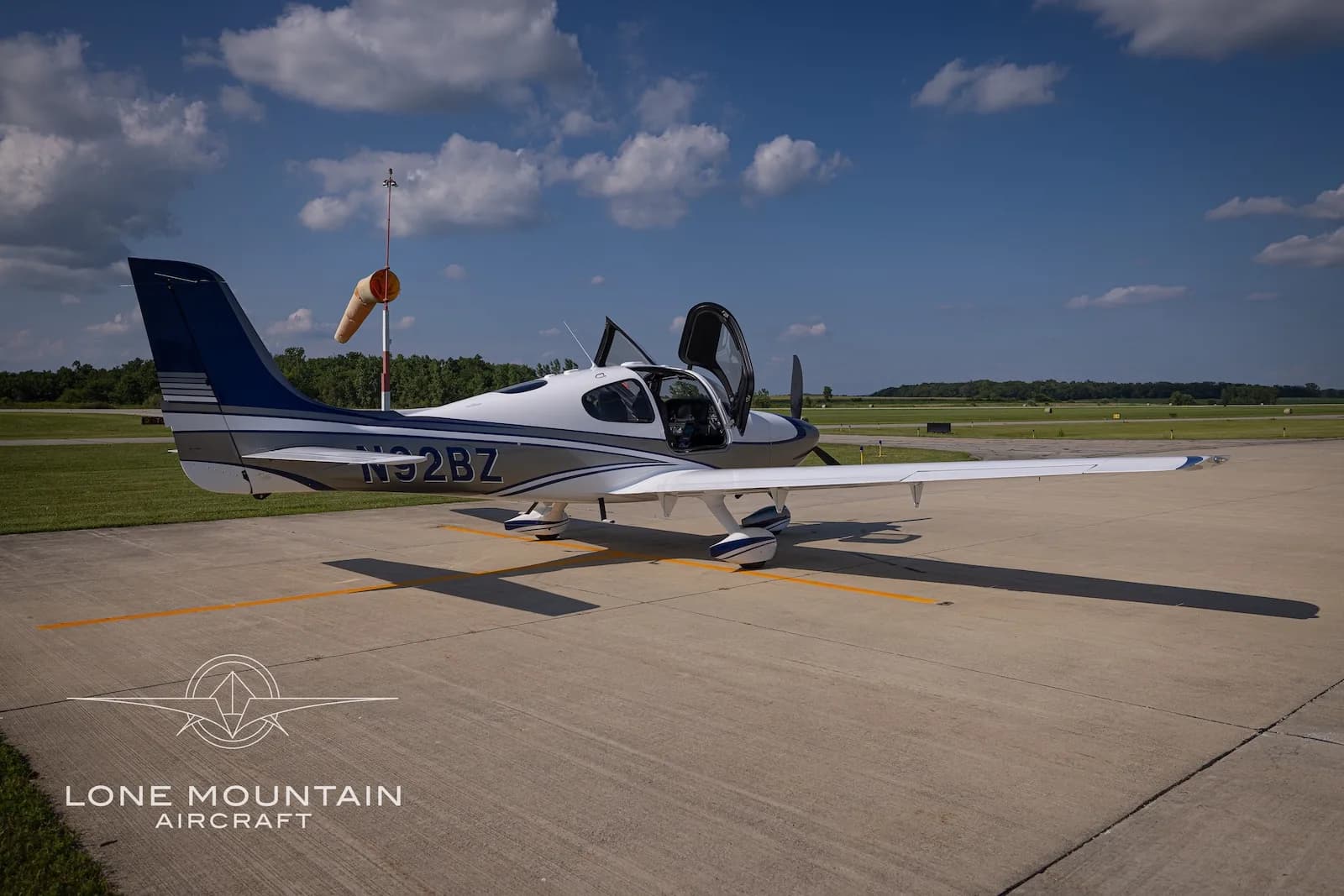 A three-quarter rear view of the white and silver Cirrus SR22 aircraft N92BZ with blue accents, parked on an airfield with its doors open.