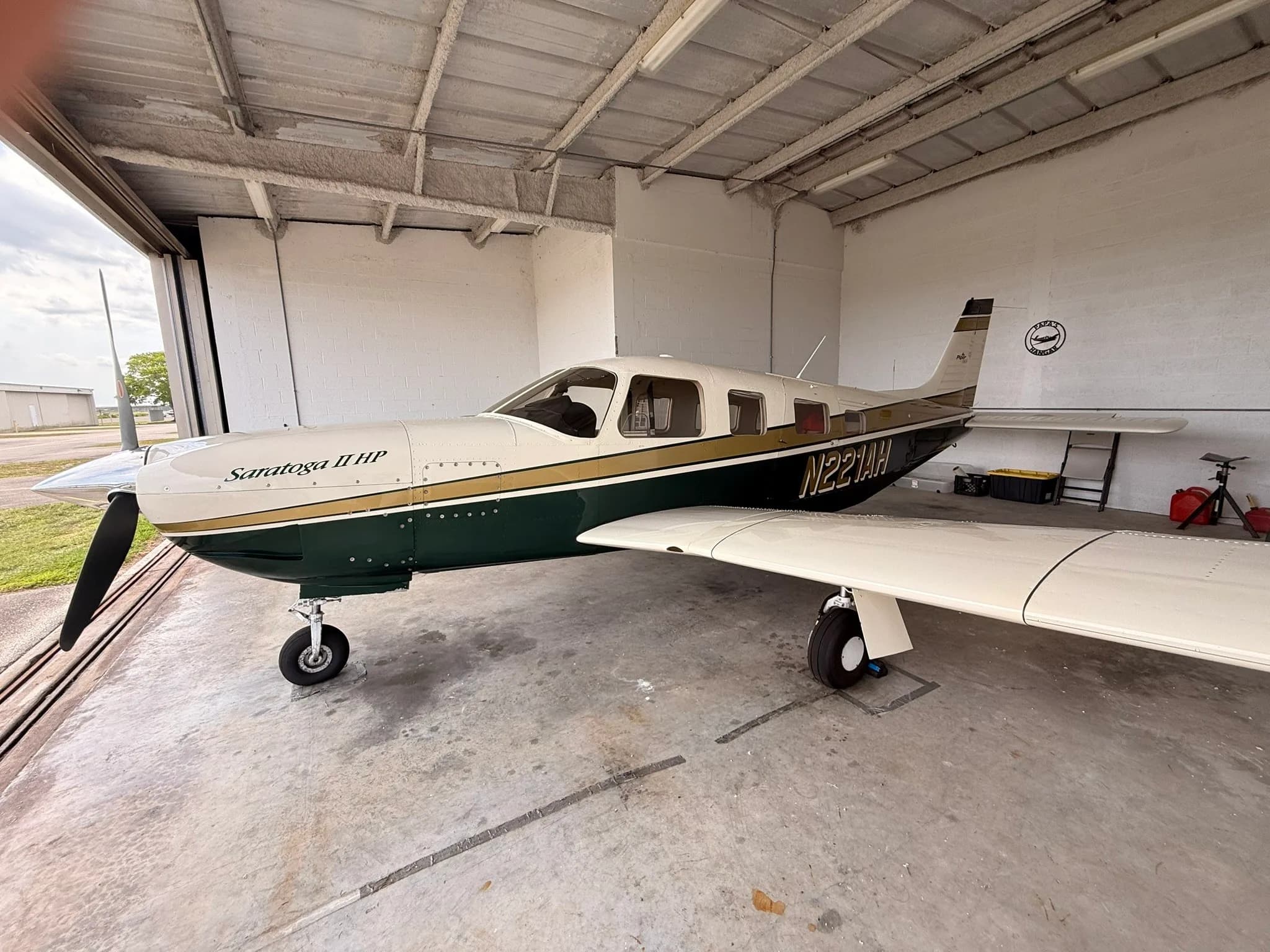 Side view of a Piper Saratoga II HP, N221AH, in a white, gold, and dark green paint scheme, parked inside a hangar.