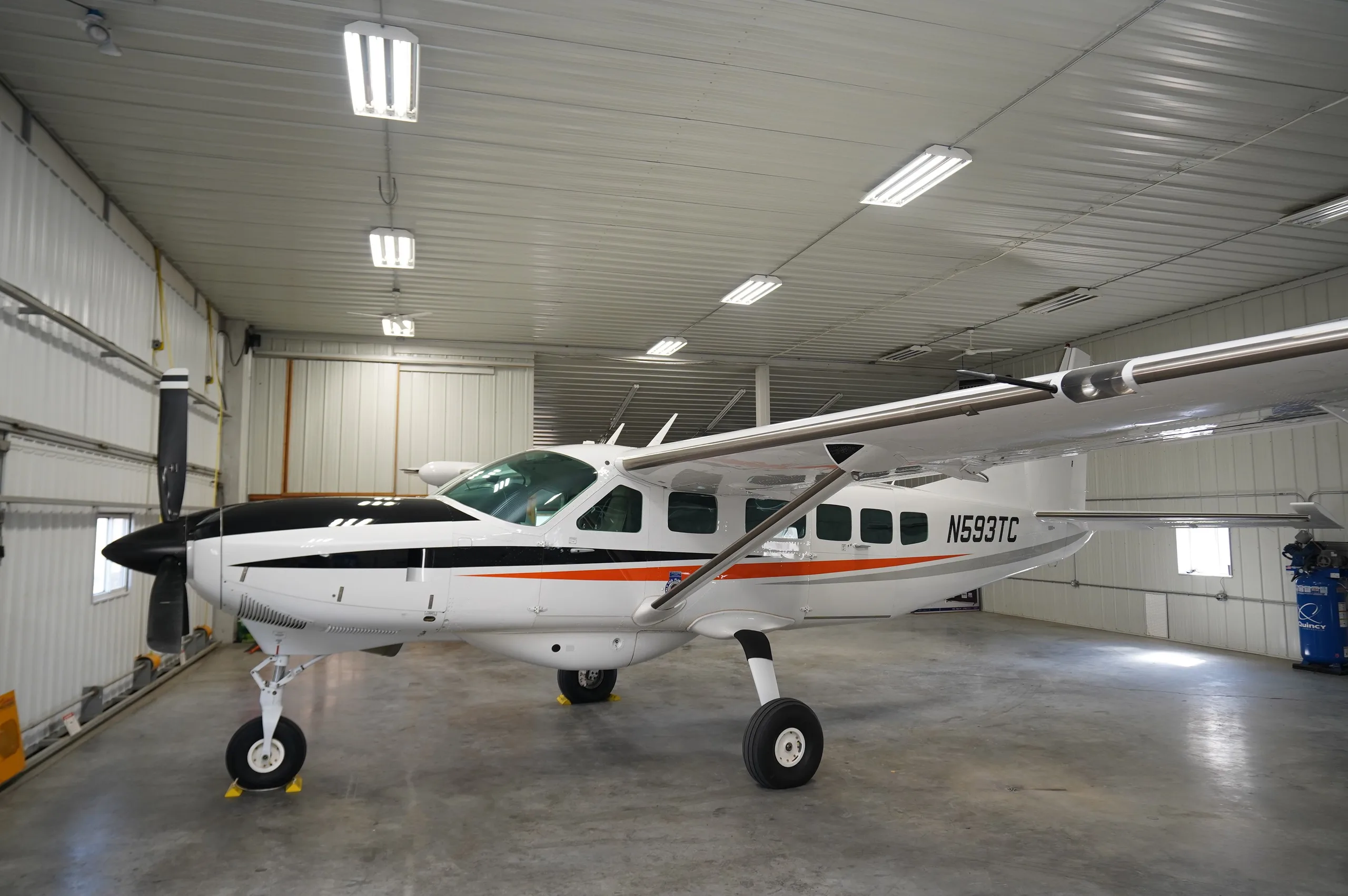 A white Cessna Caravan (N593TC) with black and orange stripes parked inside a well-lit hangar.
