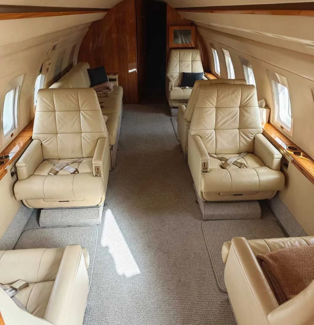 A view of the business jet's cabin, showing multiple rows of light beige leather seats and a central aisle.