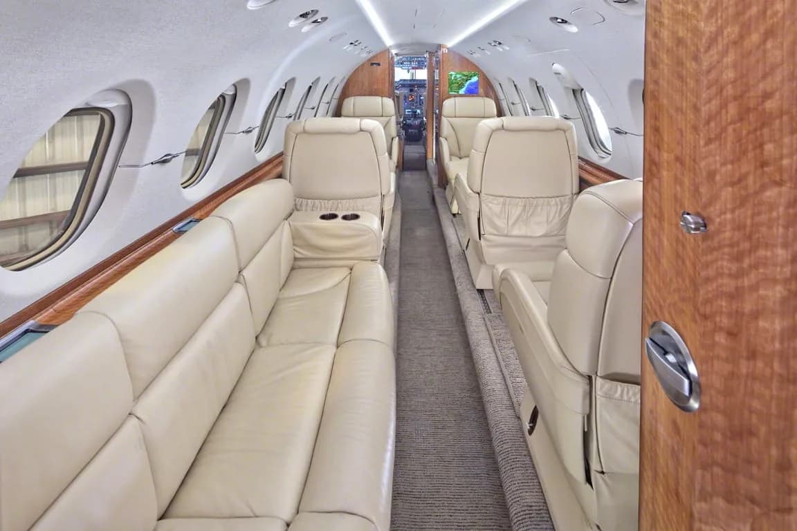 The long view of a private jet cabin with a beige leather sofa on one side and individual seats on the other.
