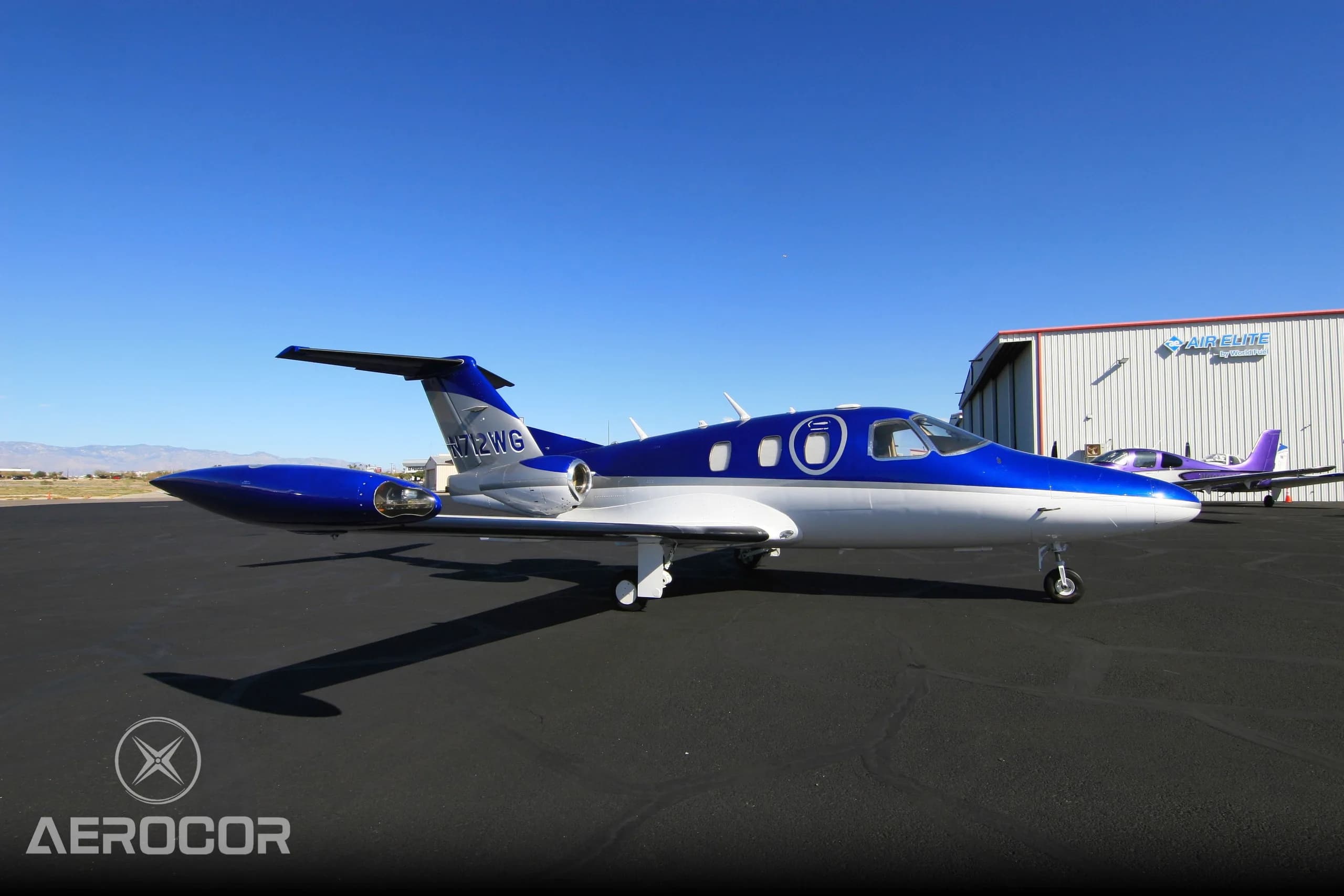 A side view of the blue and white Eclipse 500/550 jet, N712WG, on an airport tarmac with a hangar in the background.