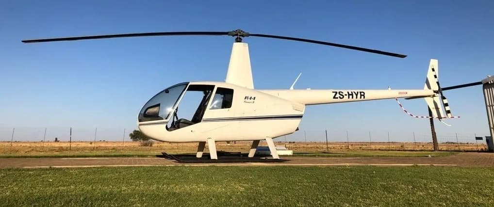 A white Robinson R44 Raven II helicopter with registration ZS-HYR is parked on a concrete pad next to a grassy field.