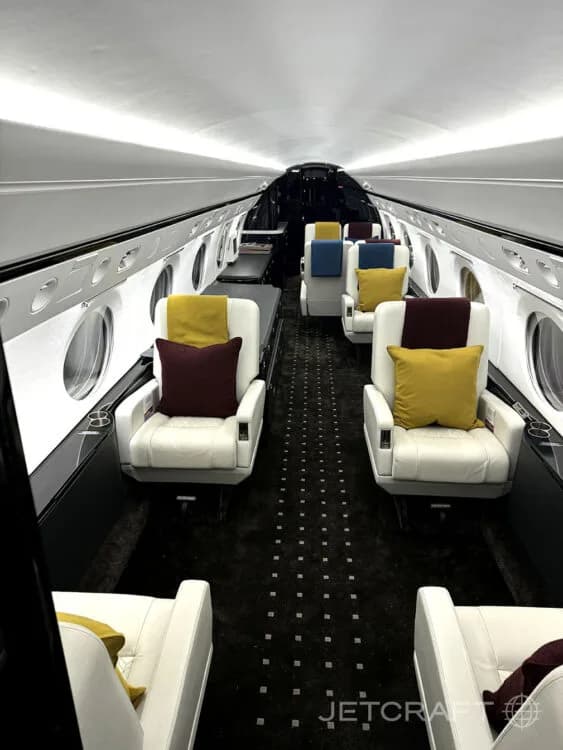 The luxurious interior cabin of a private jet, featuring white leather seats with colorful accent pillows and a dark patterned carpet.