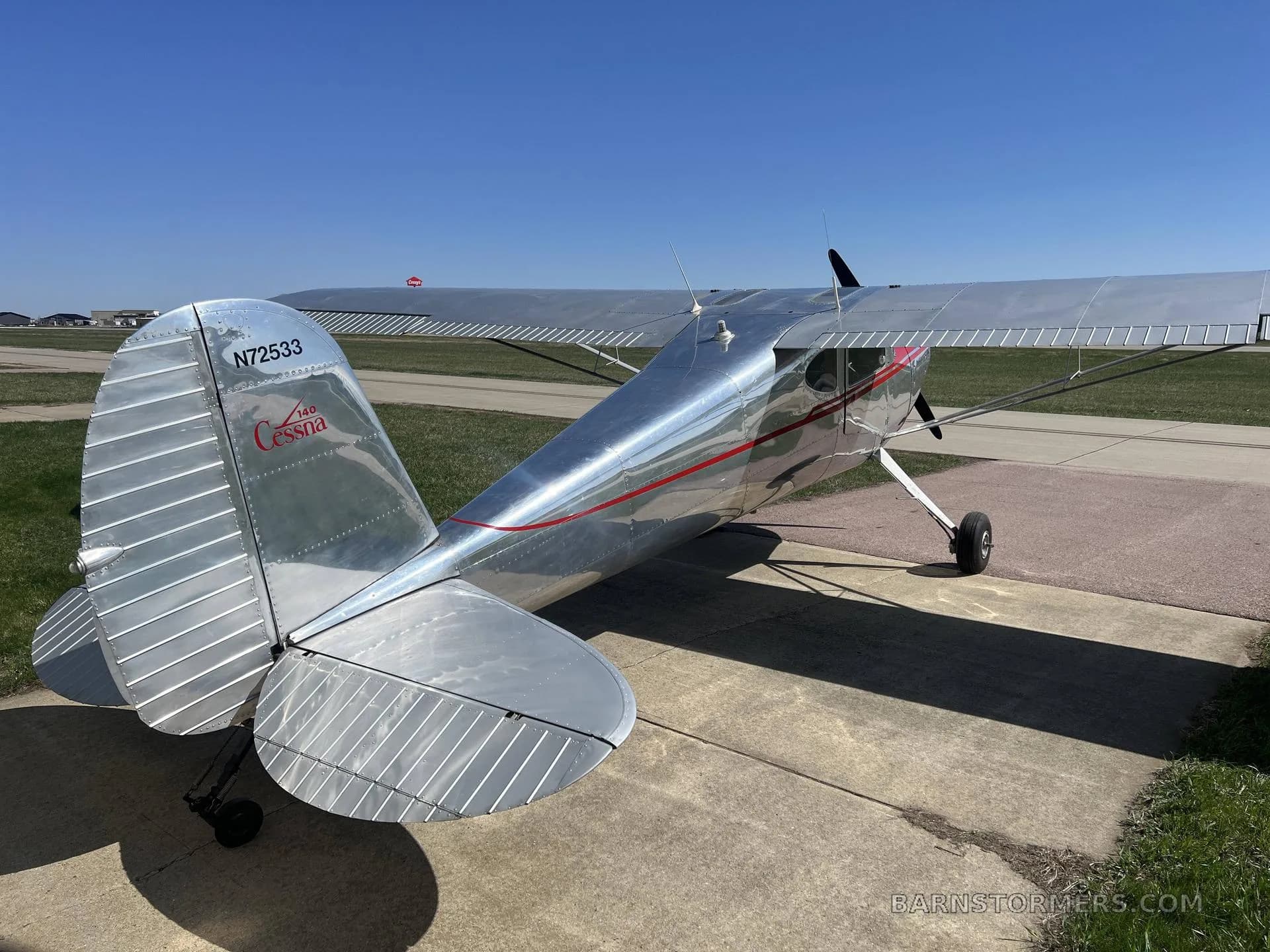 Rear-quarter view of a silver Cessna 140 with red accents, showing the tail number N72533, parked on an airfield.