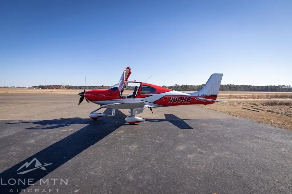 A red and silver Cirrus SR22T aircraft with open doors parked on an asphalt ramp under a clear blue sky.