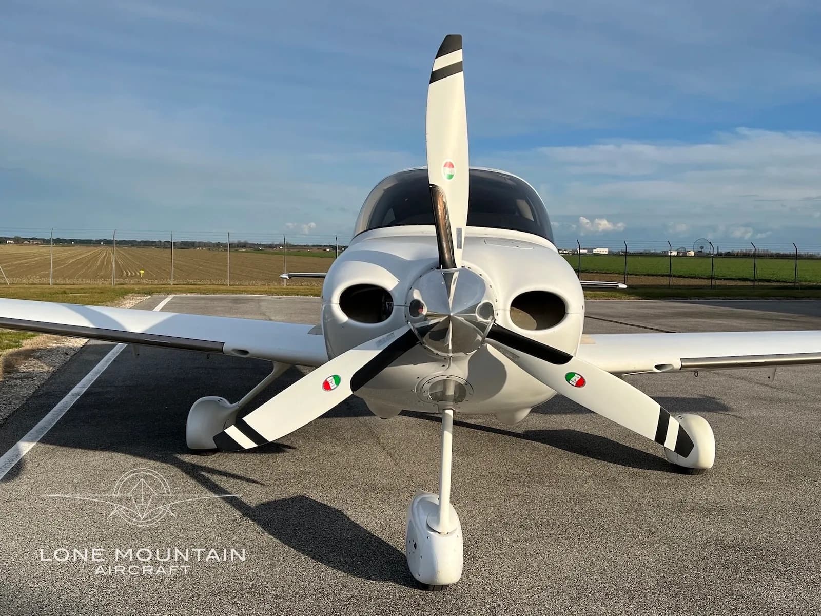 A head-on view of a white Cirrus SR22 aircraft with Italian flag colors on its propeller blades, parked on an airfield.