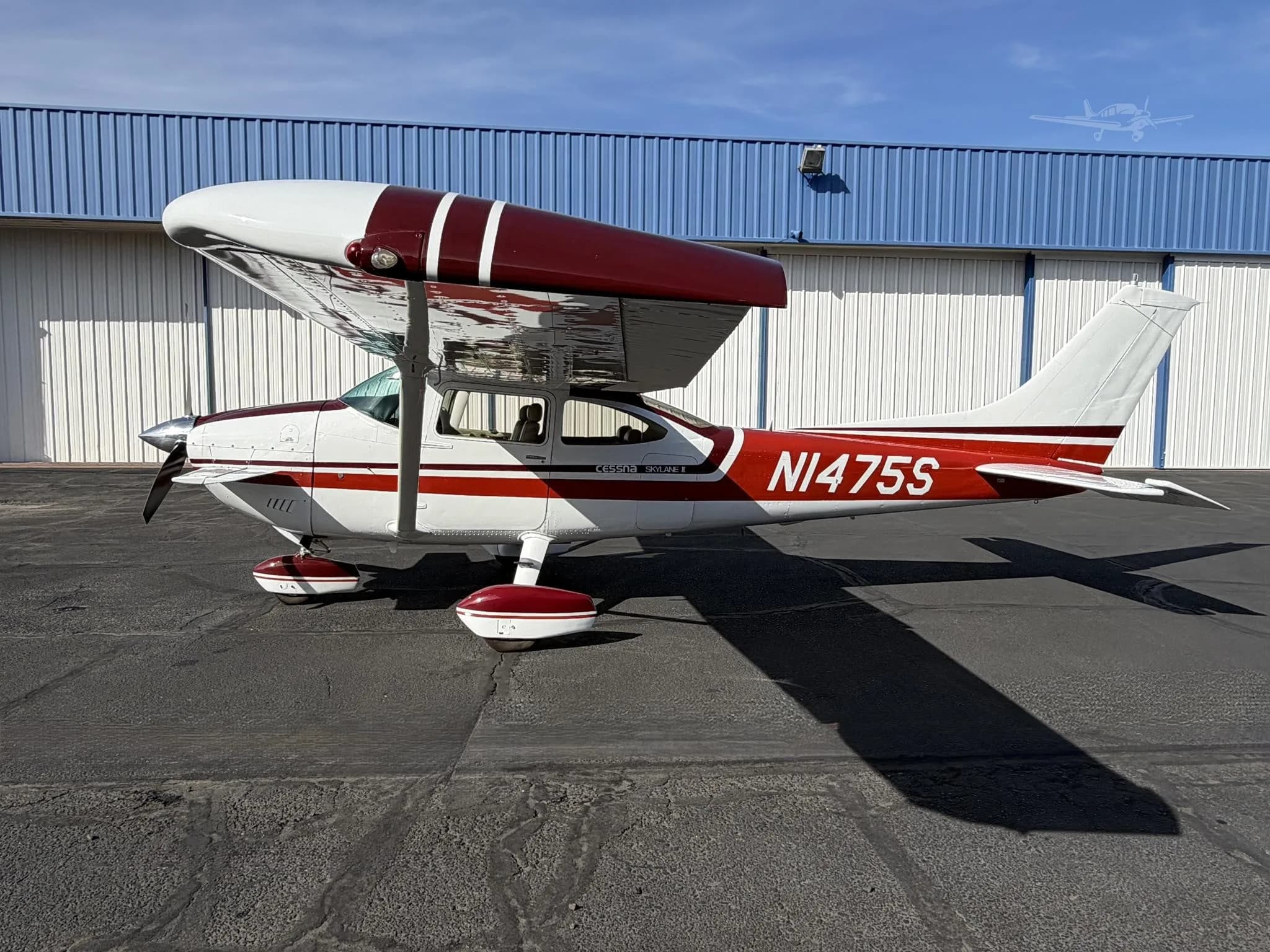 A white and red Cessna Skylane II (N1475S) parked on an asphalt apron in front of a hangar.