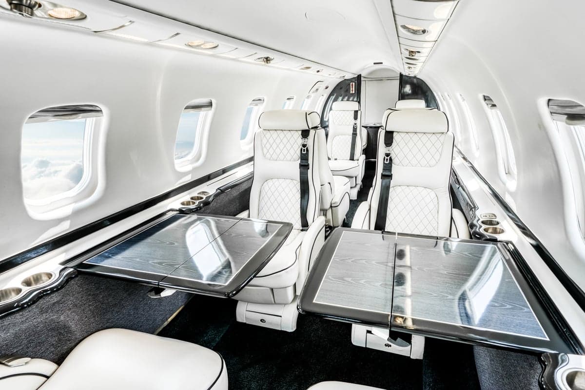 The luxurious white leather interior of a private jet cabin with two tables extended and windows showing clouds.