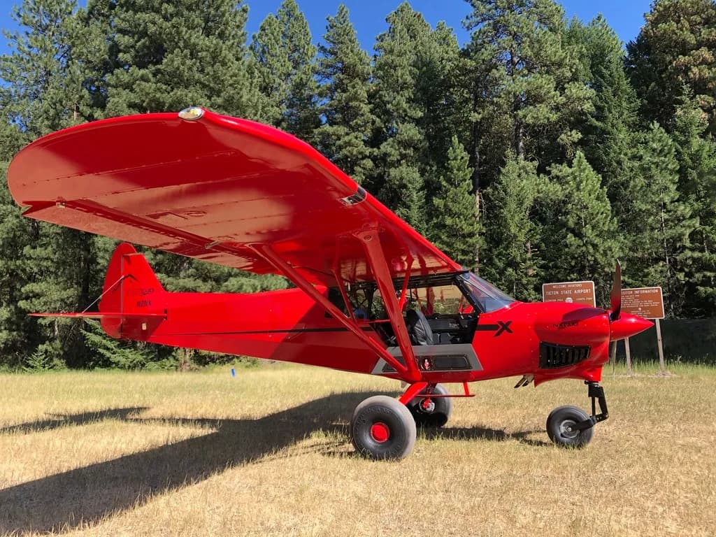 A red CubCrafters XCub parked on a grassy field with trees in the background.