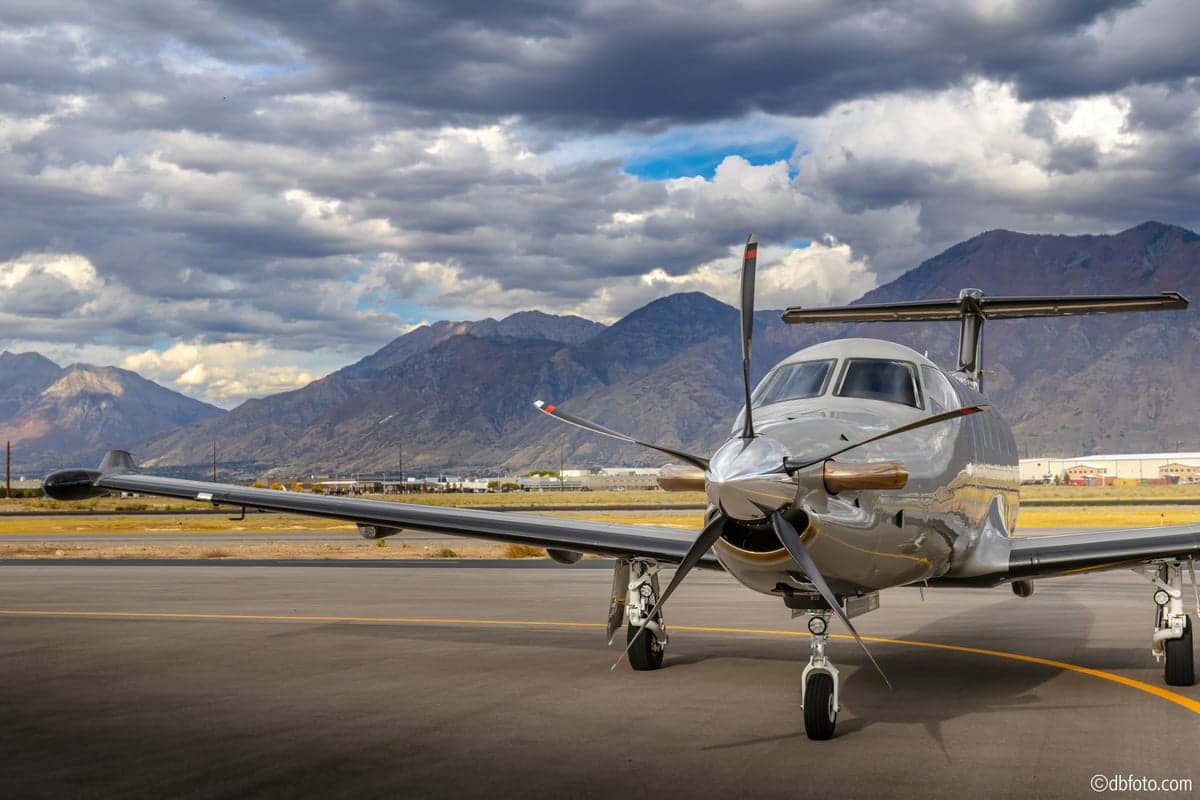 A silver Pilatus PC-12 turboprop aircraft with a black propeller parked on an airfield with mountains in the background.