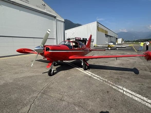 A red aircraft, HB-EML, parked on the tarmac with mountains in the background.