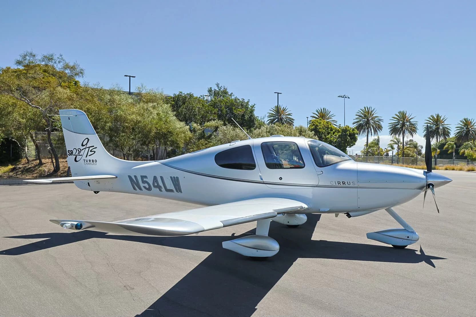 Three-quarter front view of a silver and white Cirrus SR22T (N54LW) on an tarmac with palm trees in the background.