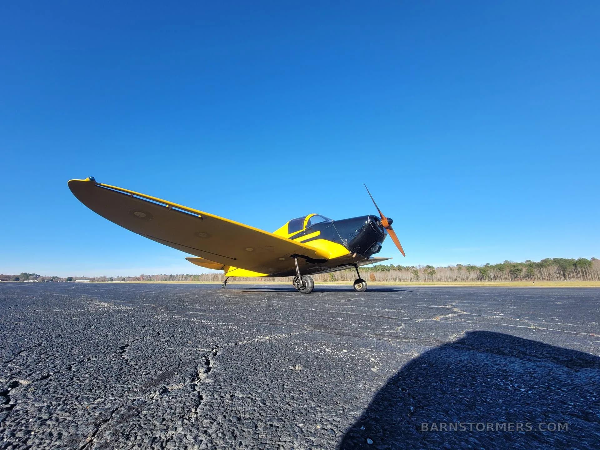 A low-angle front-right view of a yellow and black single-engine aircraft on an asphalt runway under a clear blue sky.