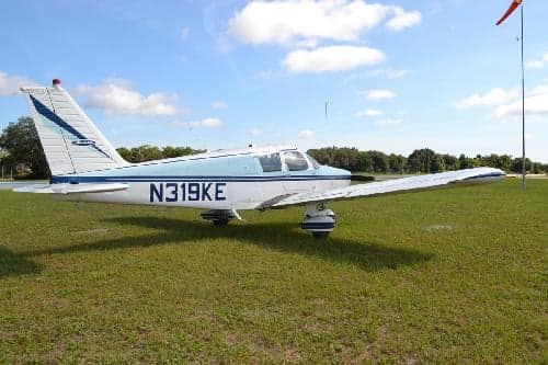 A white and blue Piper PA-28-140 Cherokee is parked on the grass with a windsock in the background.