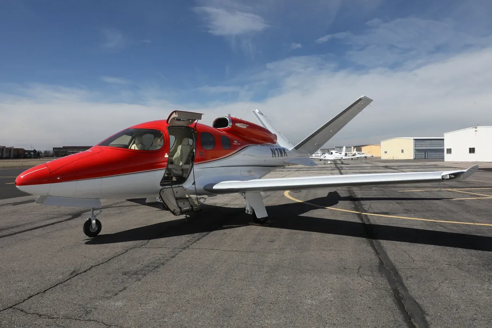 A red and white Cirrus Vision Jet (N1WK) with its main door open, parked on a tarmac, viewed from the front-left.