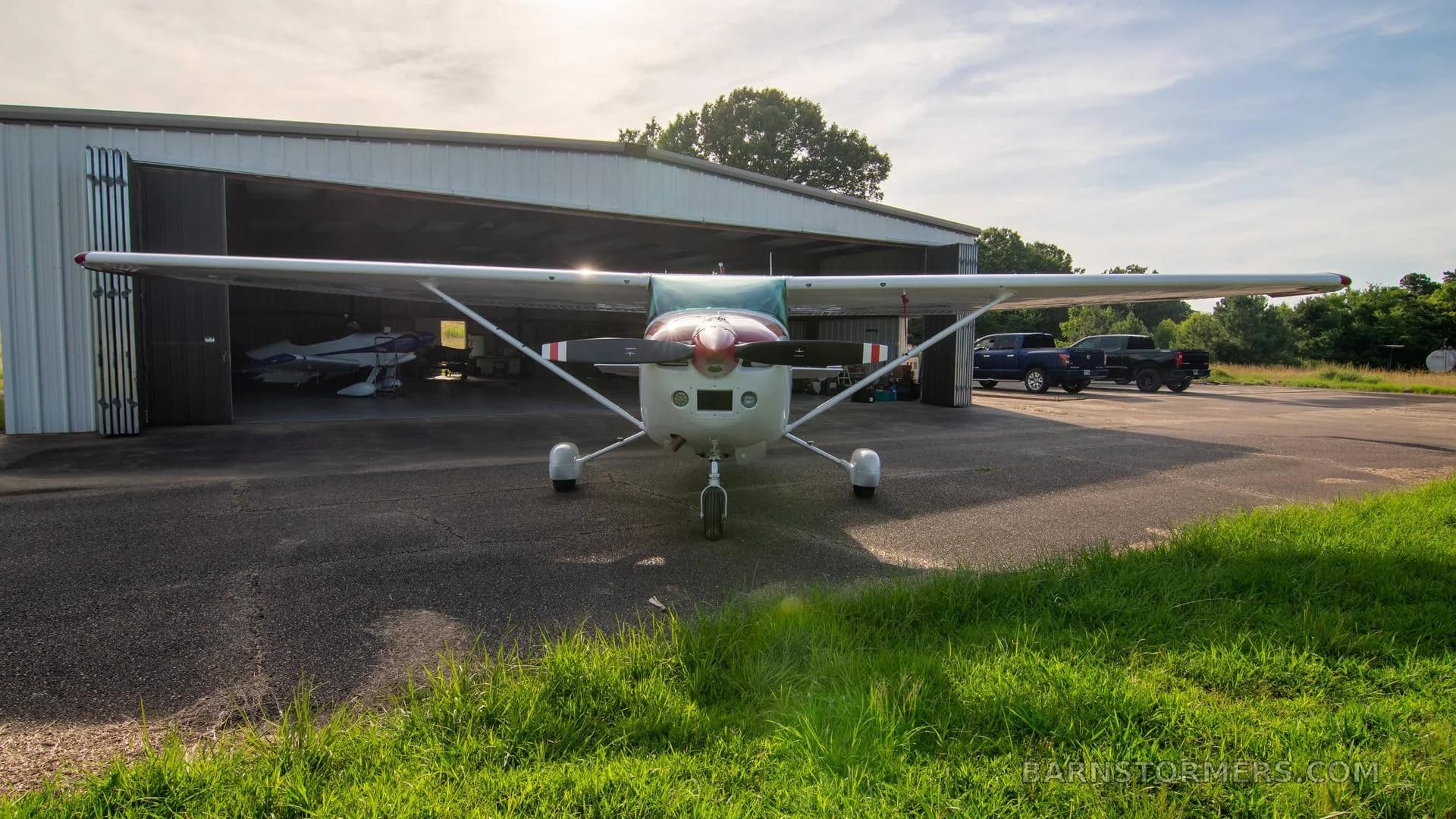 A white single-engine Cessna aircraft with red and gold stripes is viewed head-on, parked in front of an open hangar.