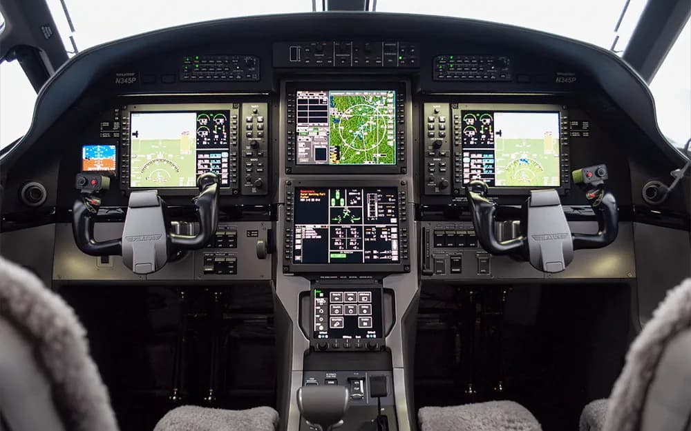 The modern glass cockpit of a Pilatus PC-12 NGX, featuring multiple large display screens and control yokes.