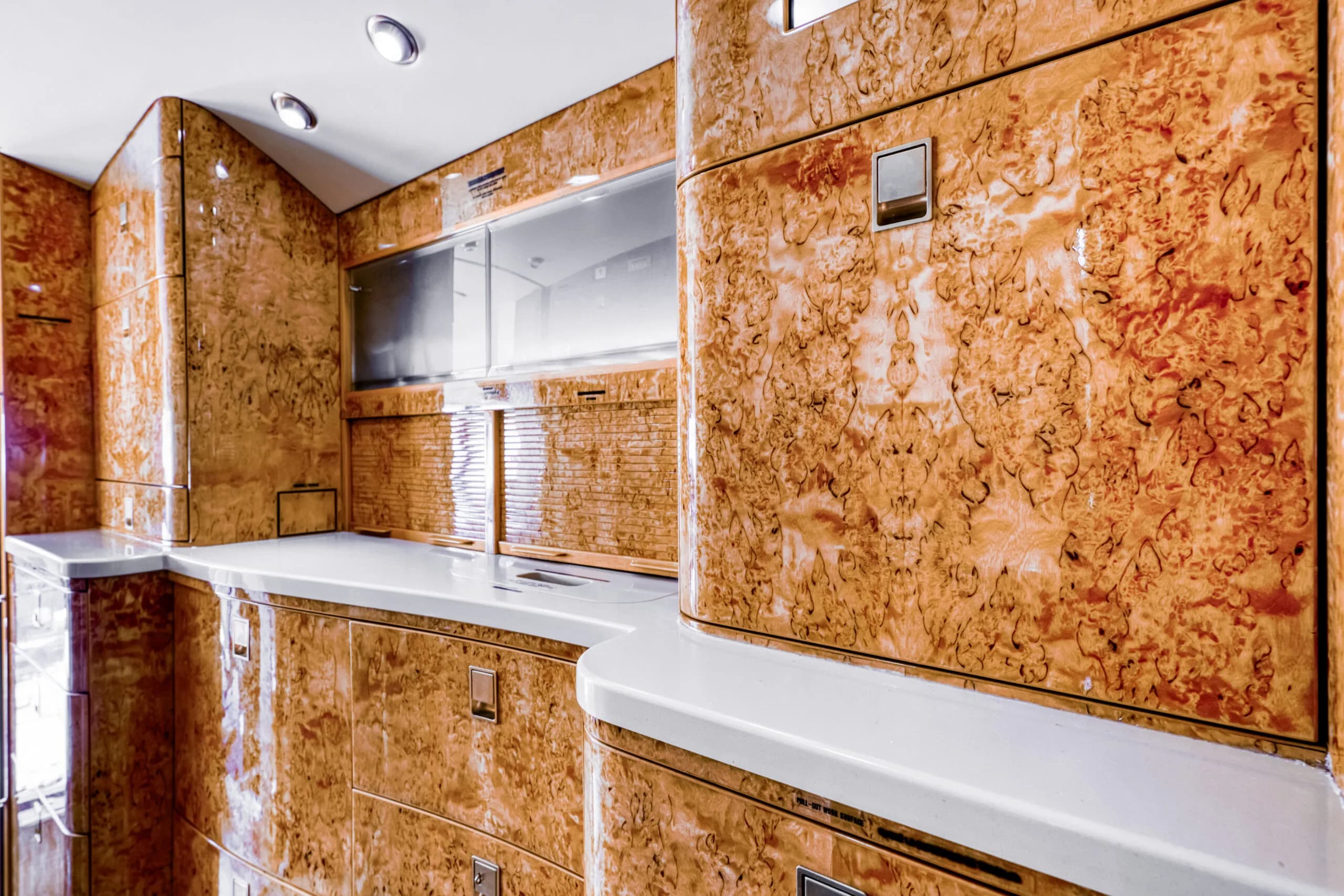 A view of the aircraft galley or refreshment center featuring light countertops and polished burled wood cabinetry.