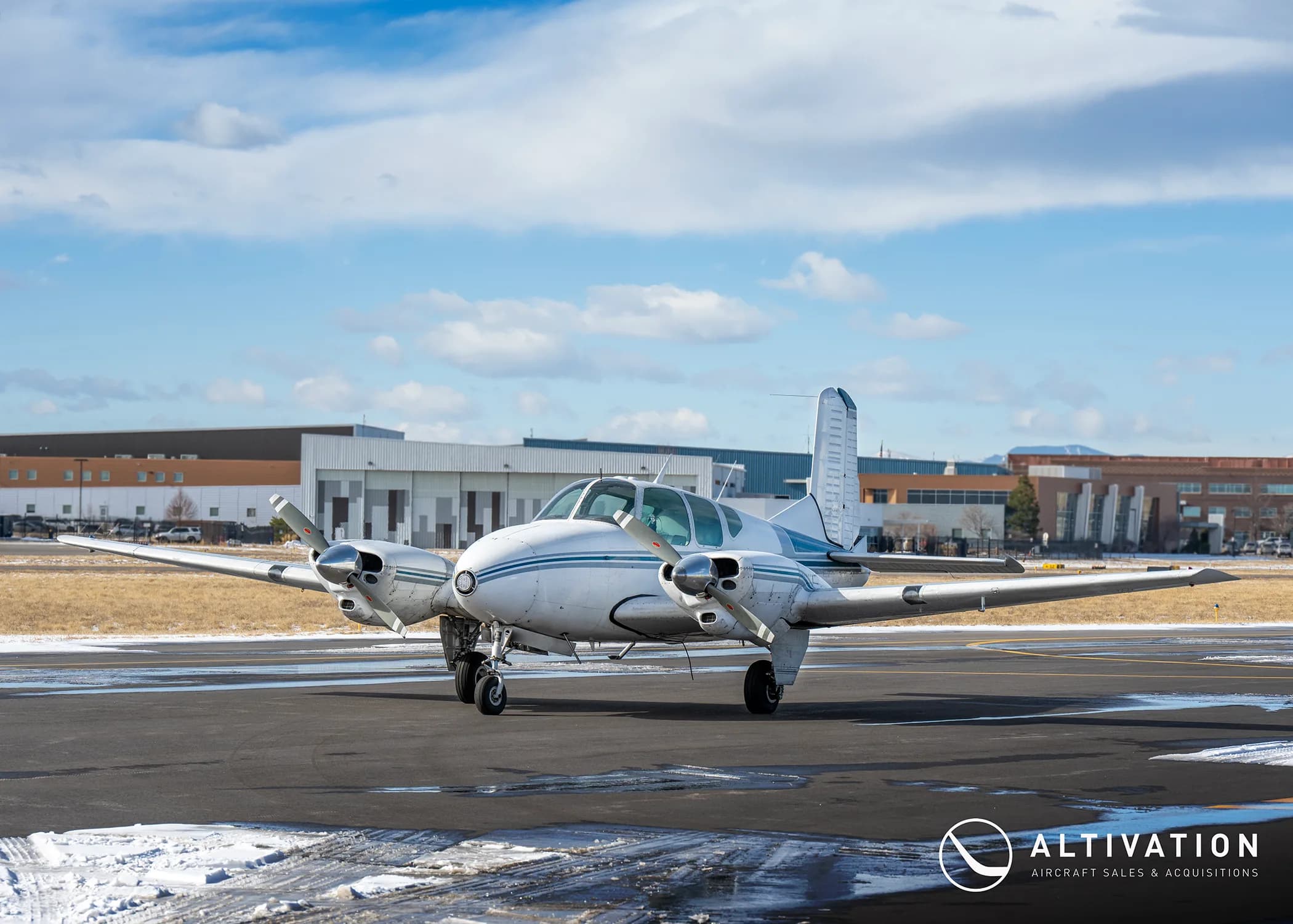 A white and blue twin-engine aircraft is parked on a wet tarmac with buildings in the background.
