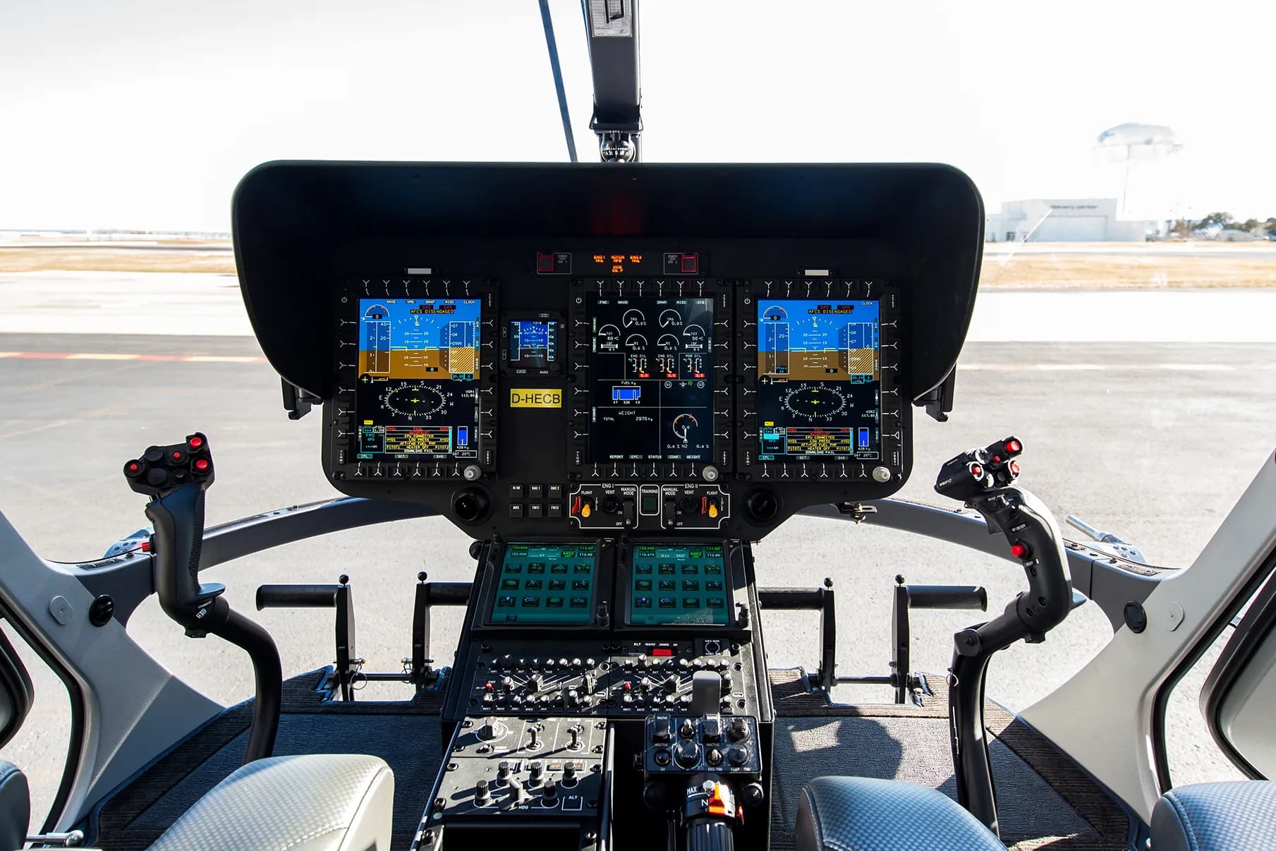 The modern glass cockpit of a helicopter with multiple digital displays and controls, overlooking an airfield.