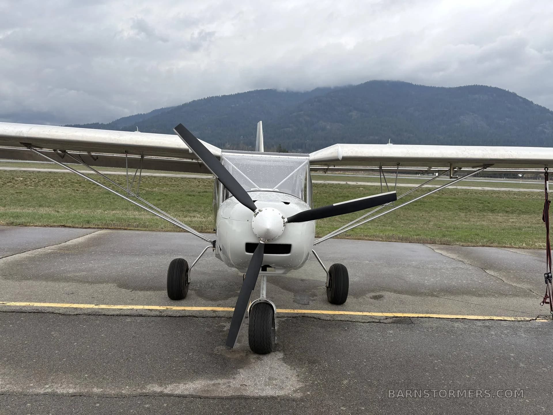 A front view of a white single-engine light aircraft with a three-blade propeller, parked on an asphalt runway under a cloudy sky.