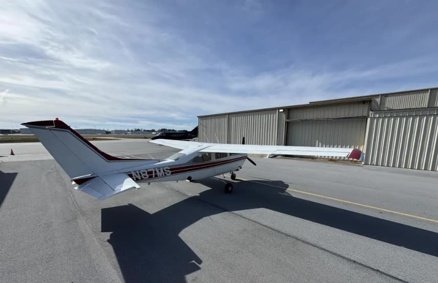 A distant rear-left view of a white and red Cessna 182 Skylane (N87MS) on an airport tarmac.