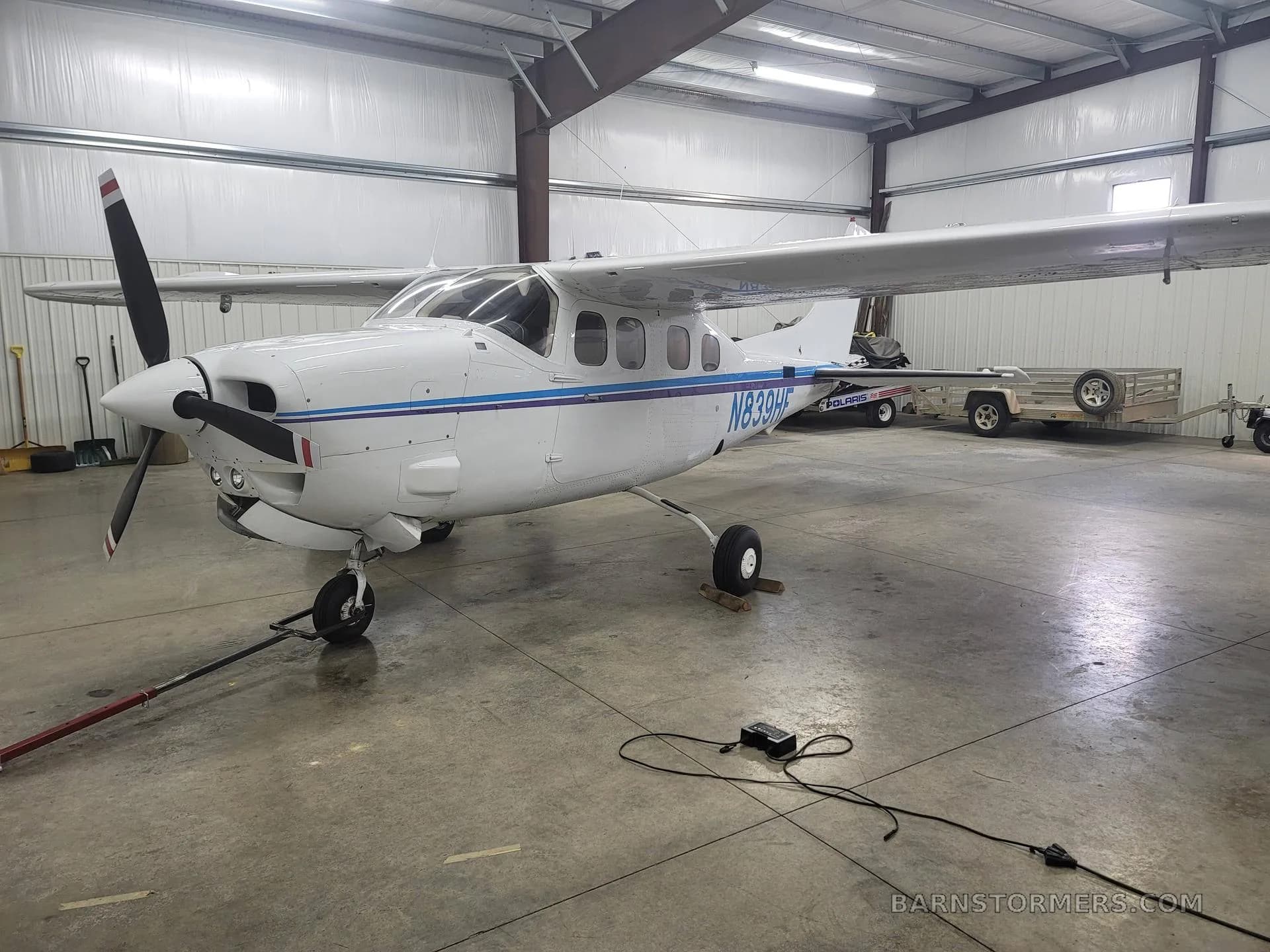 A white single-engine Cessna aircraft with blue and purple stripes, parked in a hangar.