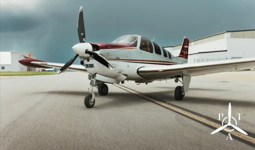 A white and red single-engine propeller aircraft, a Beechcraft Bonanza, parked on an asphalt runway under a cloudy sky.