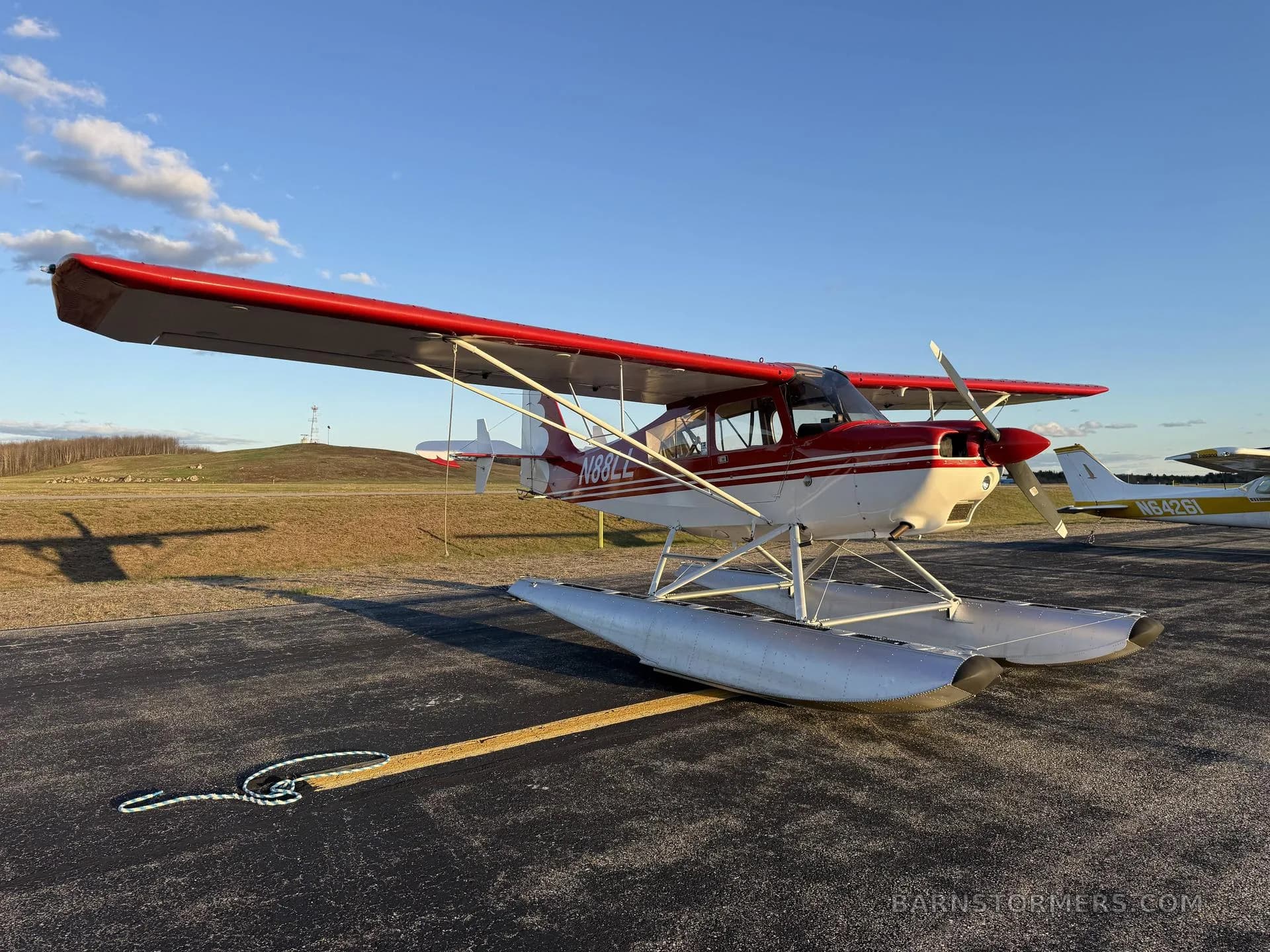 A red and white floatplane with tail number N88LL, parked on an asphalt runway with another aircraft in the background.