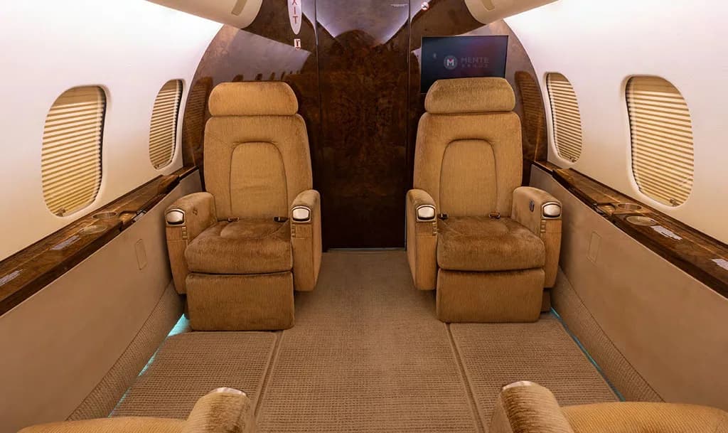 The luxurious interior cabin of a private jet, featuring two plush tan individual seats facing forward and dark wood accents.