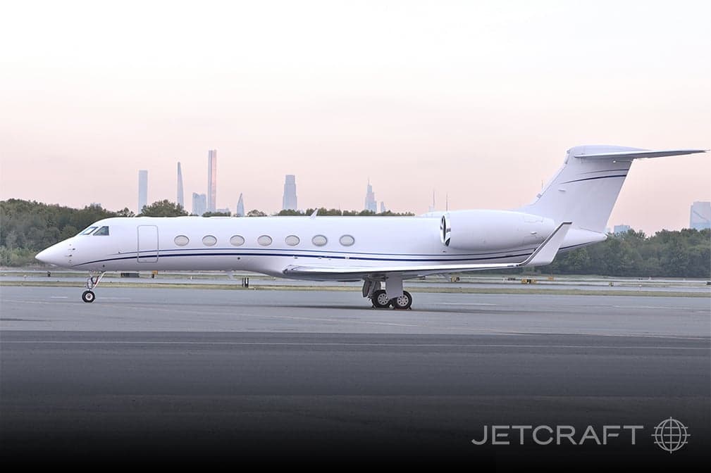 A white Gulfstream G550 business jet with blue stripes parked on a tarmac with a distant city skyline.