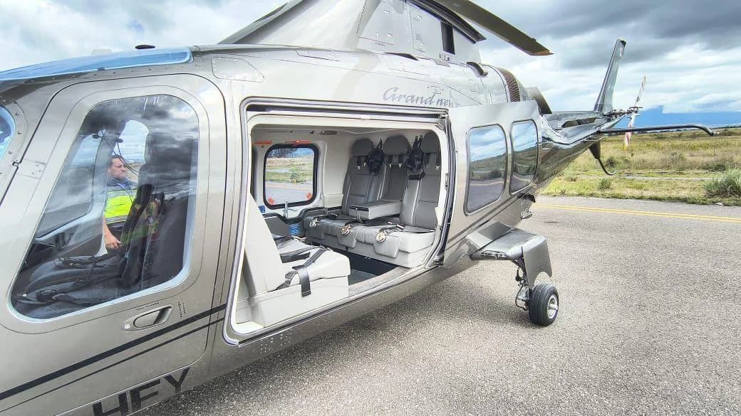 A close-up view of a grey AgustaWestland AW109 GrandNew helicopter with its passenger door open, revealing the cabin seats and a pilot in the cockpit.