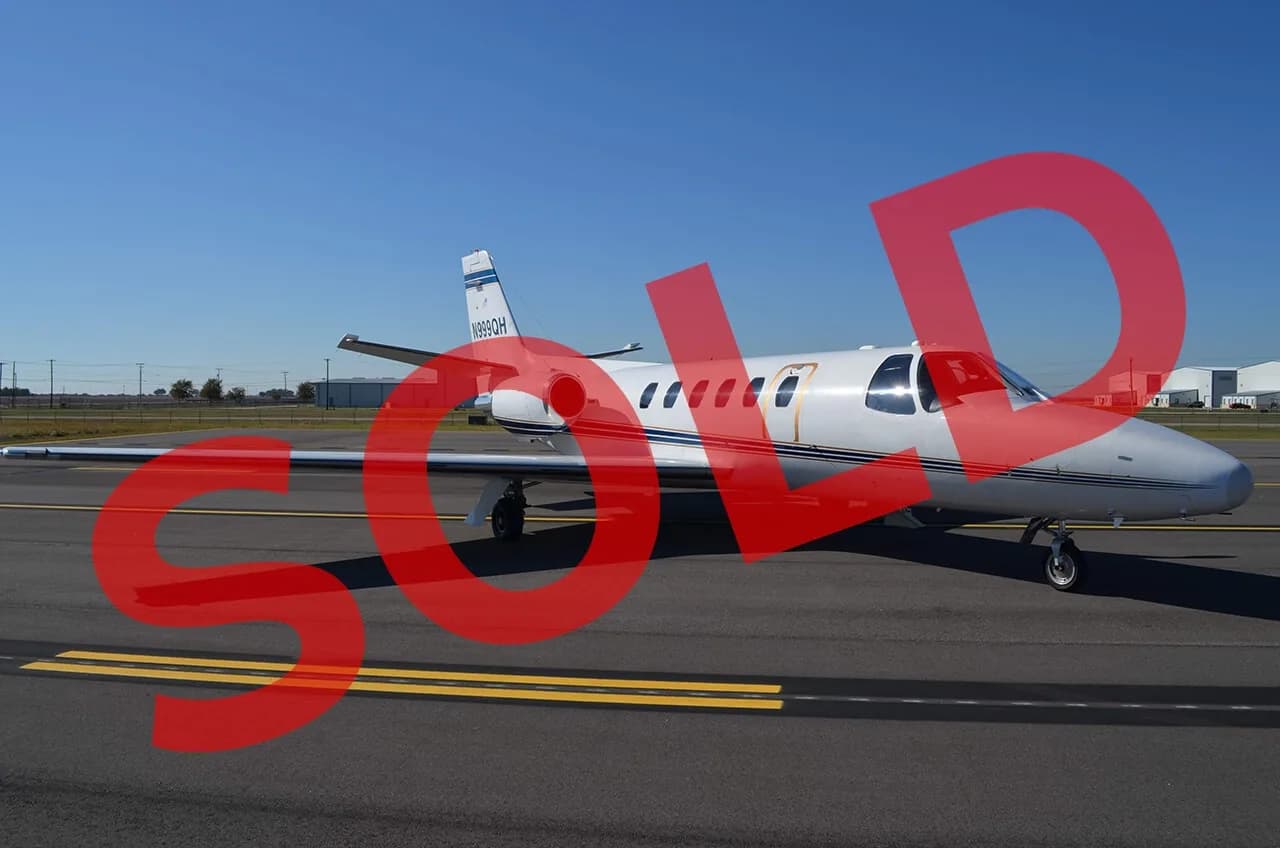 A white Cessna Citation business jet with blue and gold stripes is parked on an airport tarmac under a clear blue sky.