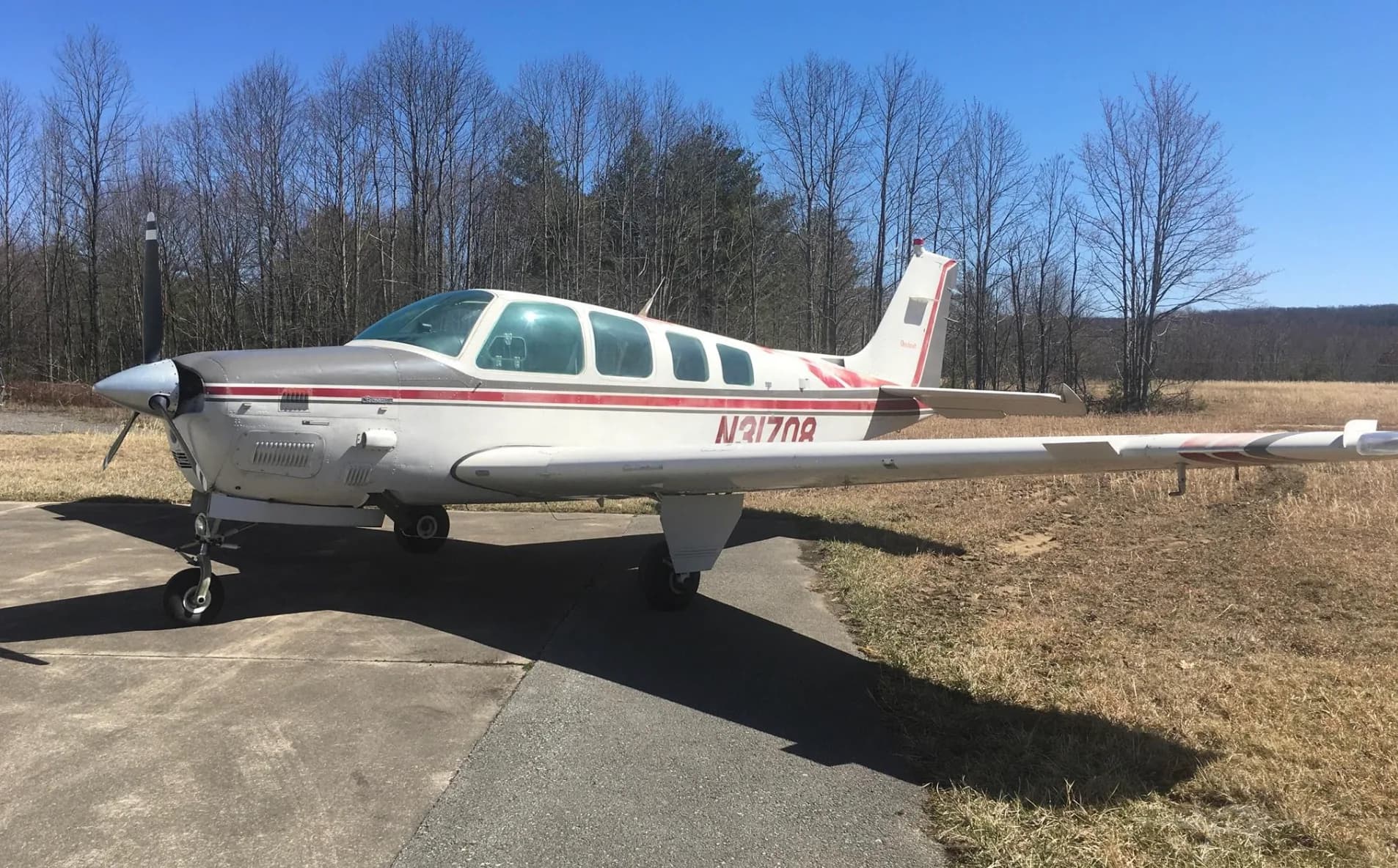 A front-quarter view of a white and red Beechcraft Bonanza N31708 parked on an asphalt ramp with bare trees in the background.