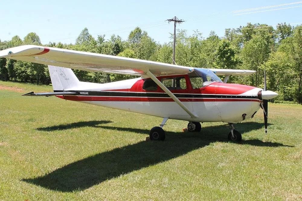A side view of a red and white Cessna 172 (N6924X) with a black stripe, parked on a sunny grassy field.
