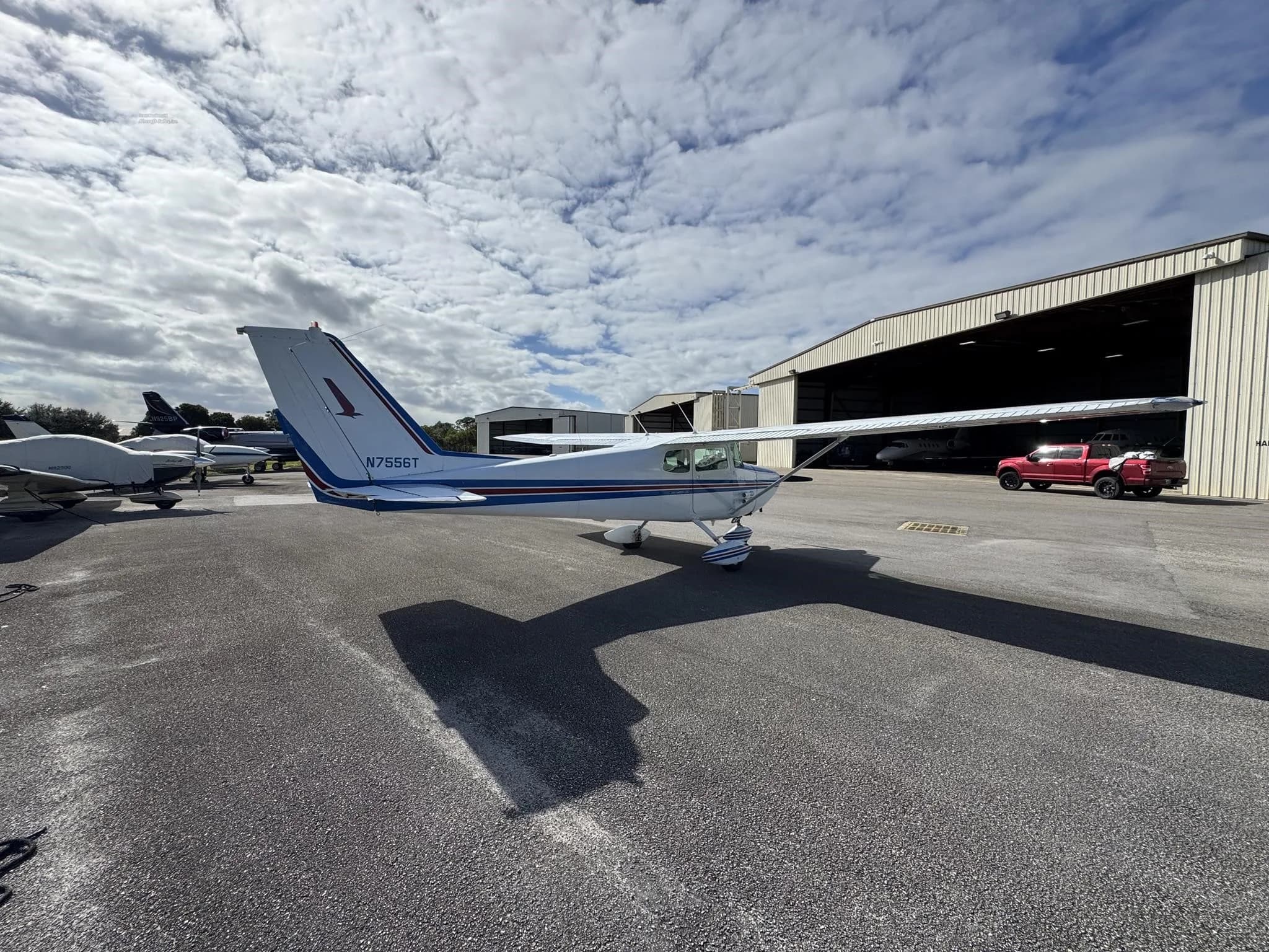 Rear-side view of a white and blue Cessna 172 (N7556T) parked on an airfield with hangars.