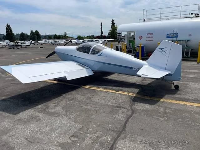 A light blue and white single-engine aircraft with tail number N7EL, parked next to a large fuel tank on an airfield tarmac.