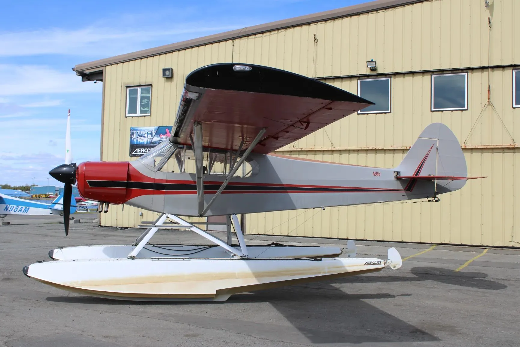 A side view of the red and grey floatplane with N984 on the tail, parked next to a yellow hangar.
