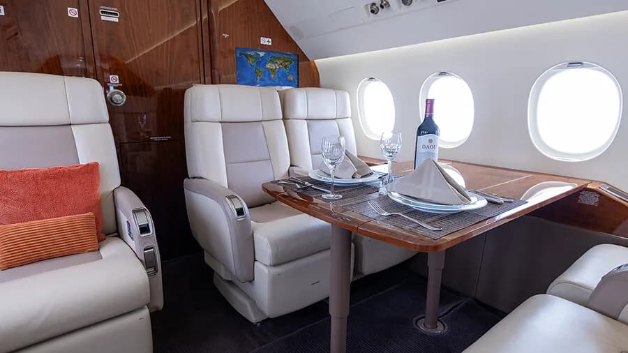 A close-up of the private jet's dining area with two beige leather seats, a table set with wine and place settings, and a world map on the bulkhead.