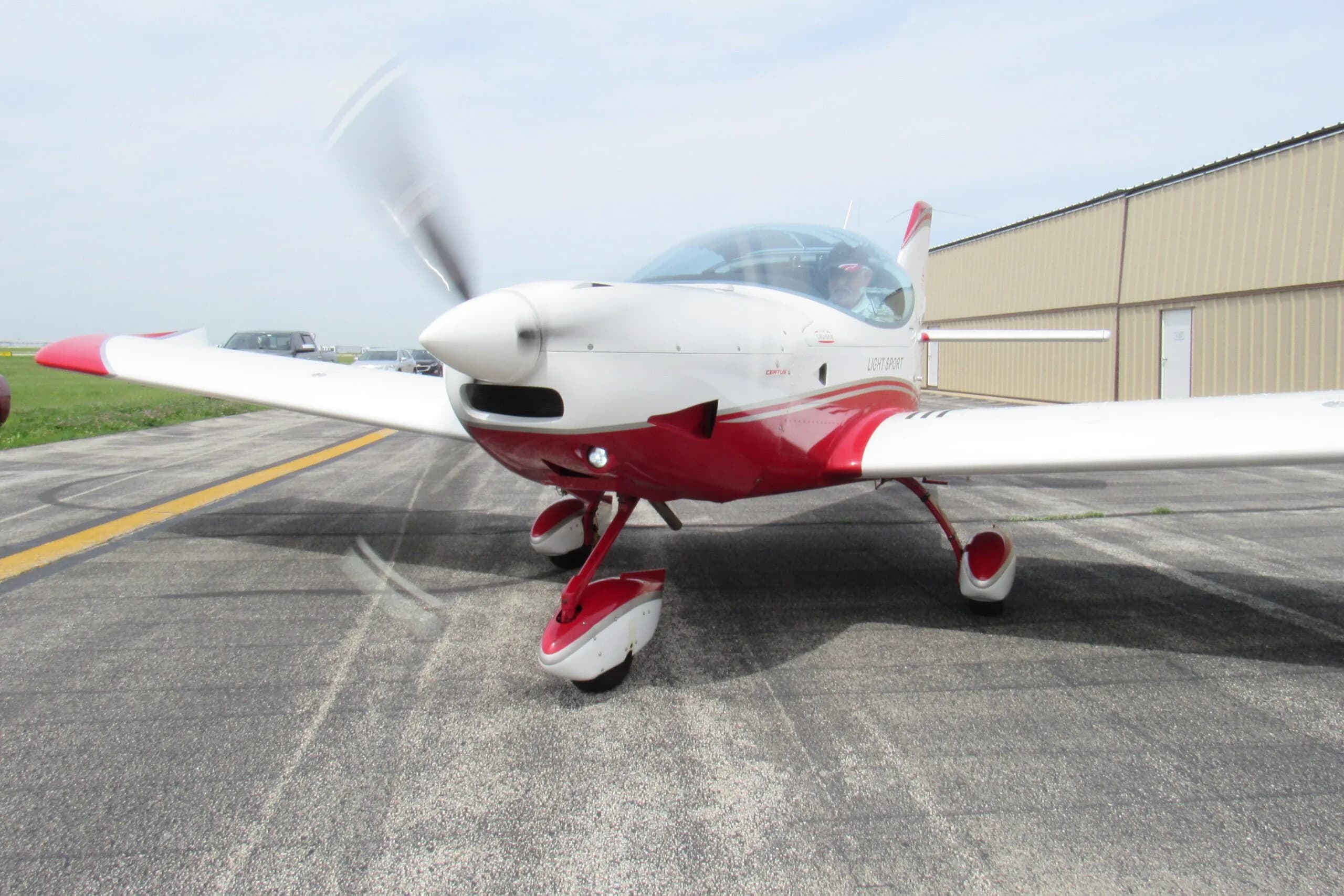 A white and red SportCruiser aircraft with a spinning propeller and a person in the cockpit on an airport tarmac.