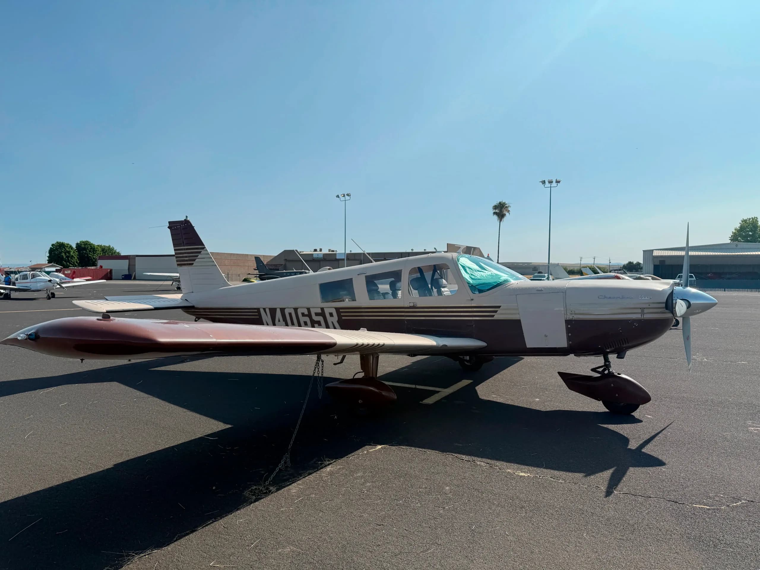 A brown and white Piper Cherokee Six aircraft, registration N4065R, is parked on an asphalt apron at an airport on a sunny day.